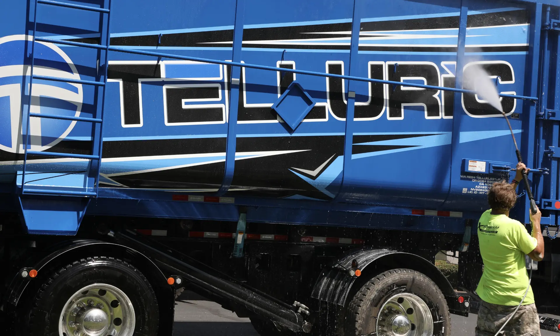 Technician in a yellow shirt spraying water while washing a blue semi trailer