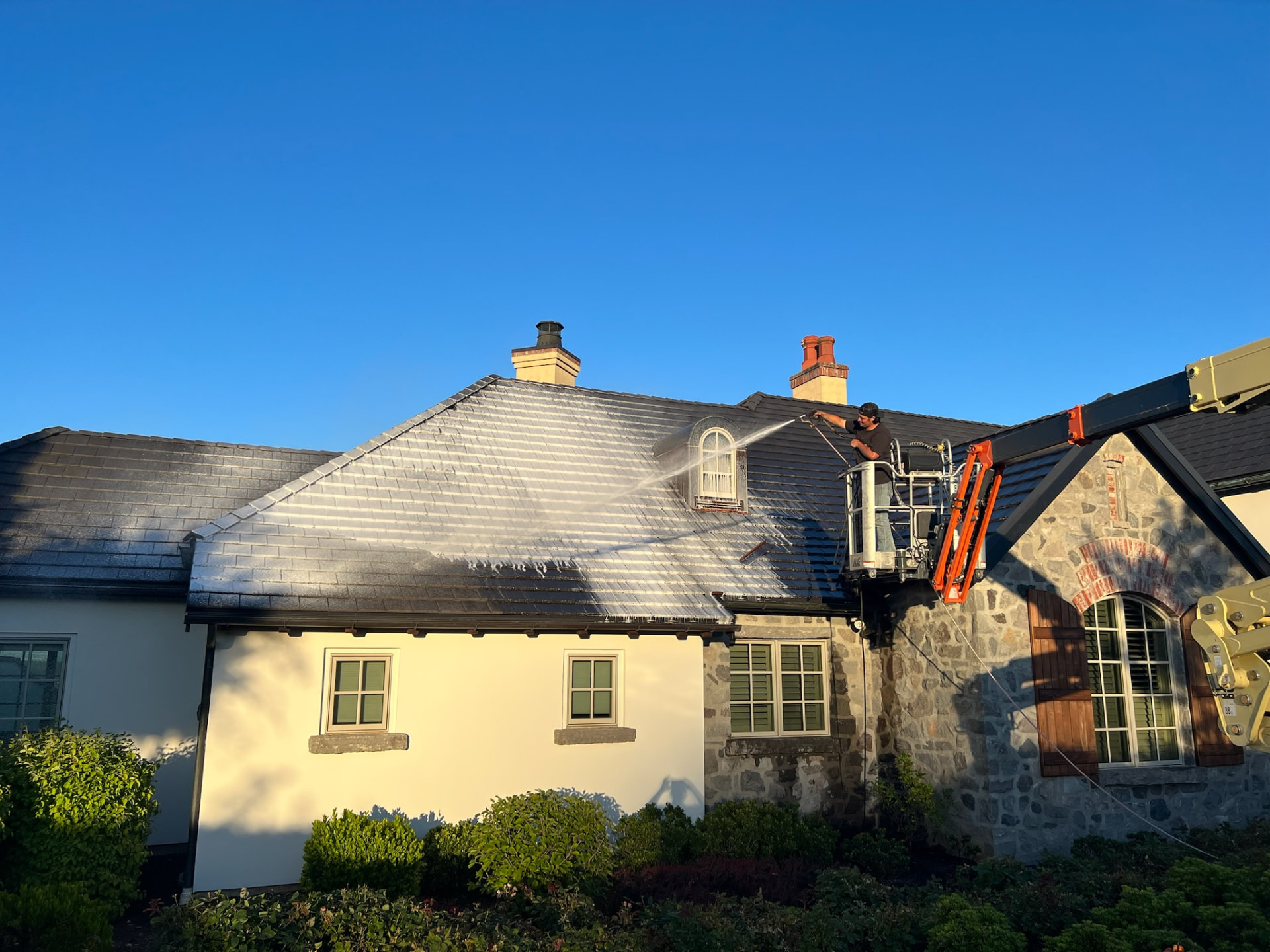 Technician pressure washing a white and stone house from a lift